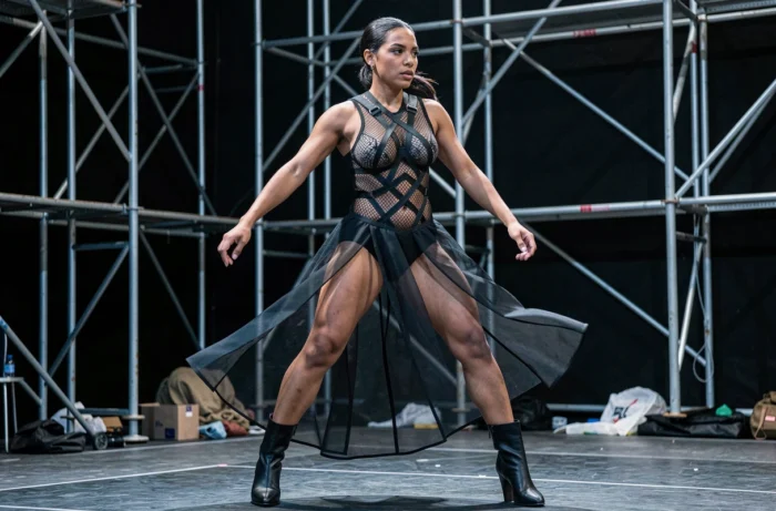 Femme en justaucorps en mesh structuré avec harnais géométrique sur scène industrielle, illustrant l'univers performatif et vestimentaire de FKA Twigs
